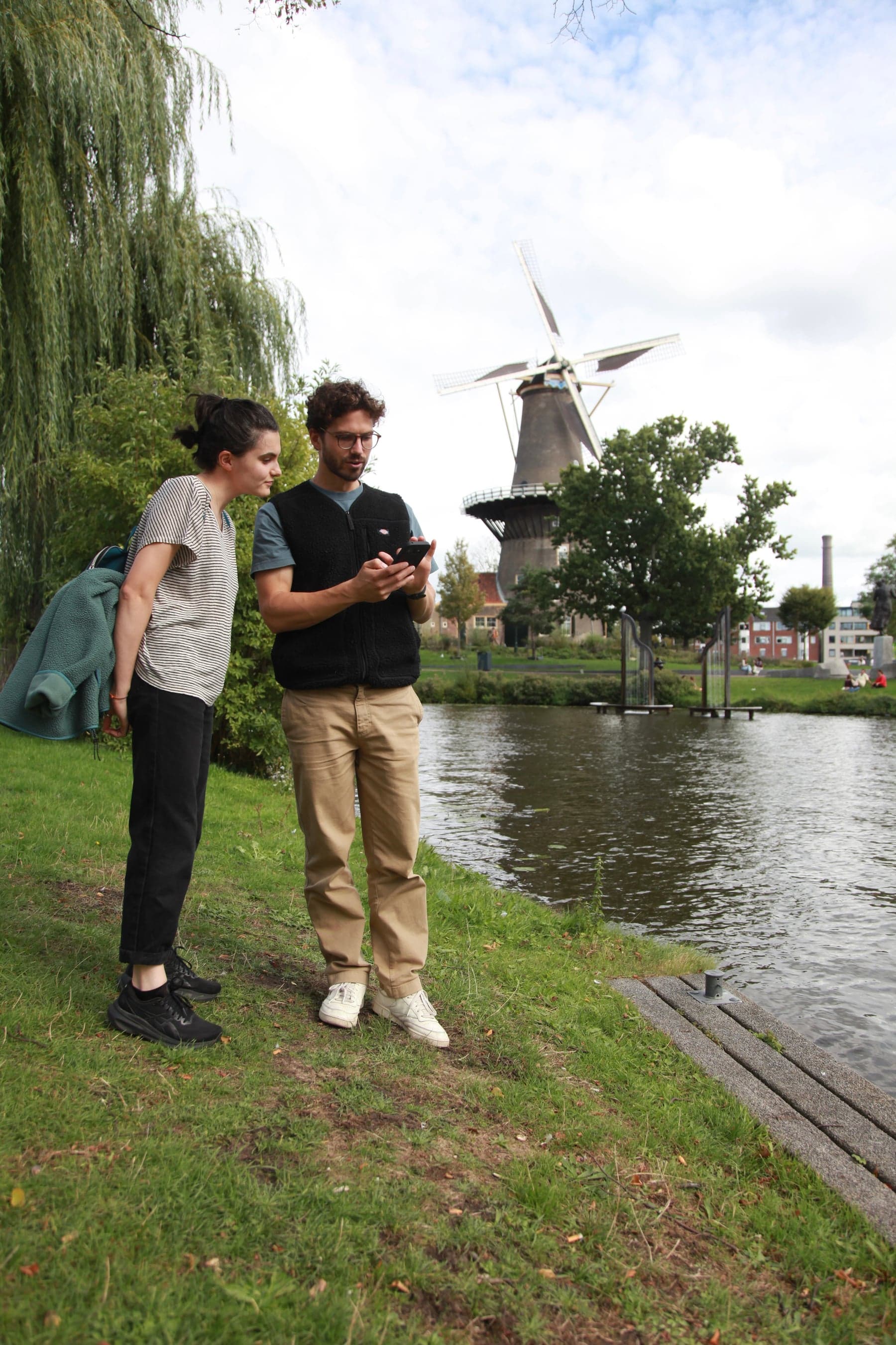 Two people reviewing a smartphone photo beside the water and a Leiden windmill